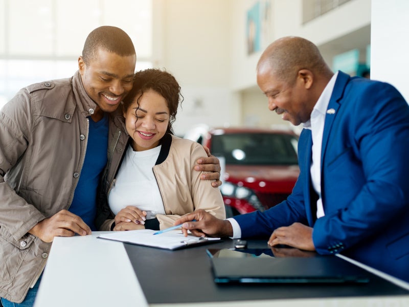 A happy couple reviews documents with a salesman in a car showroom. The man in a blue jacket smiles while pointing at a paper on the counter.
